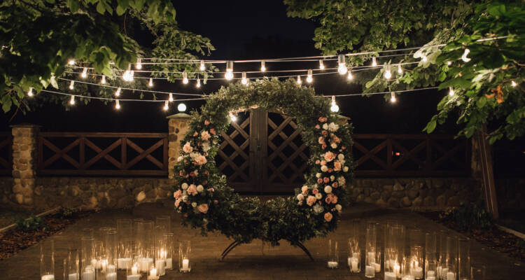 Beautiful photozone with big wreath decorated with greenery and roses in centerpiece, candles on the sides, and garland hanged between trees